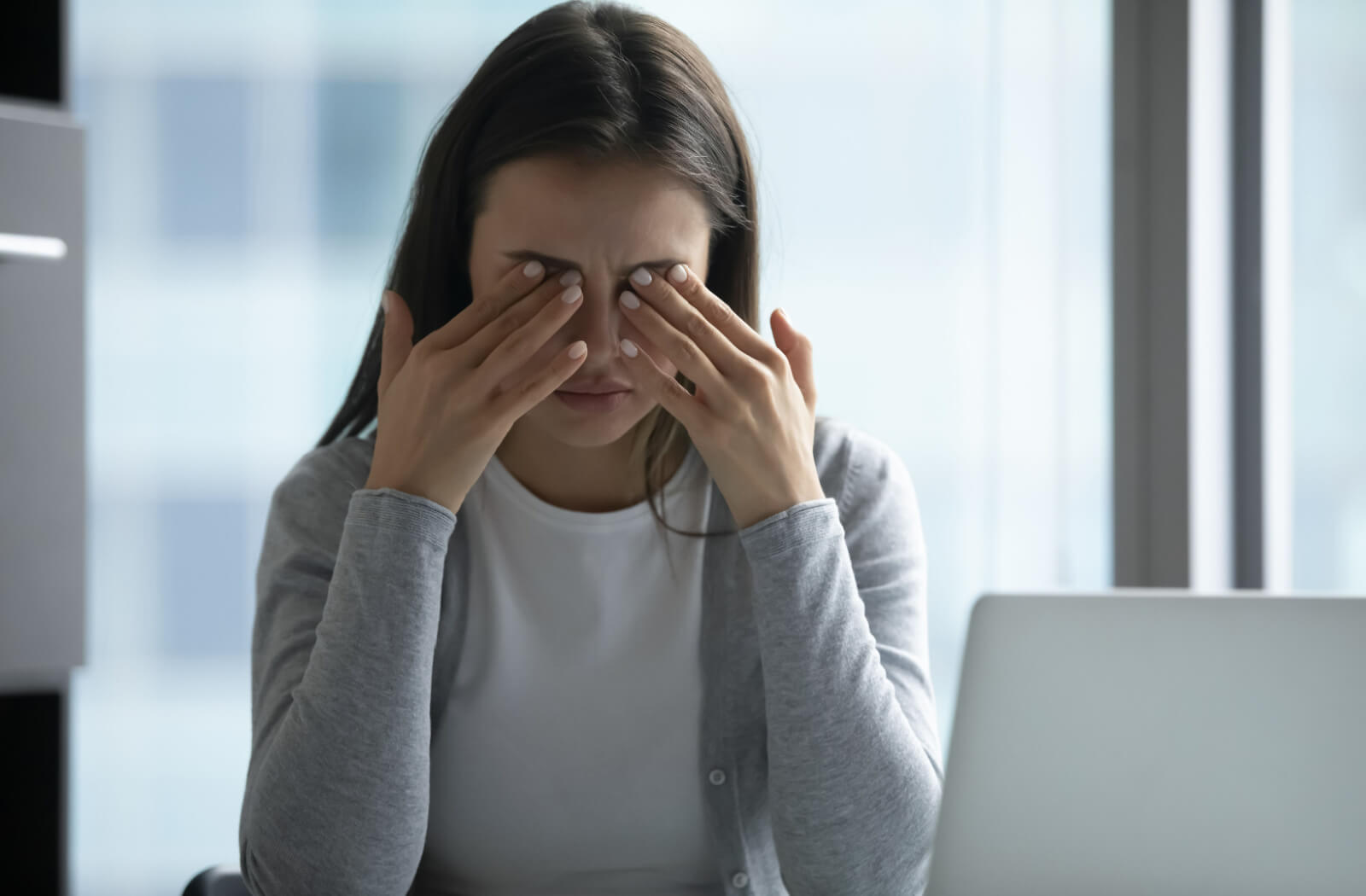 An adult working at their office in front of a laptop and rubbing their eyes in frustration due to digital eye strain.