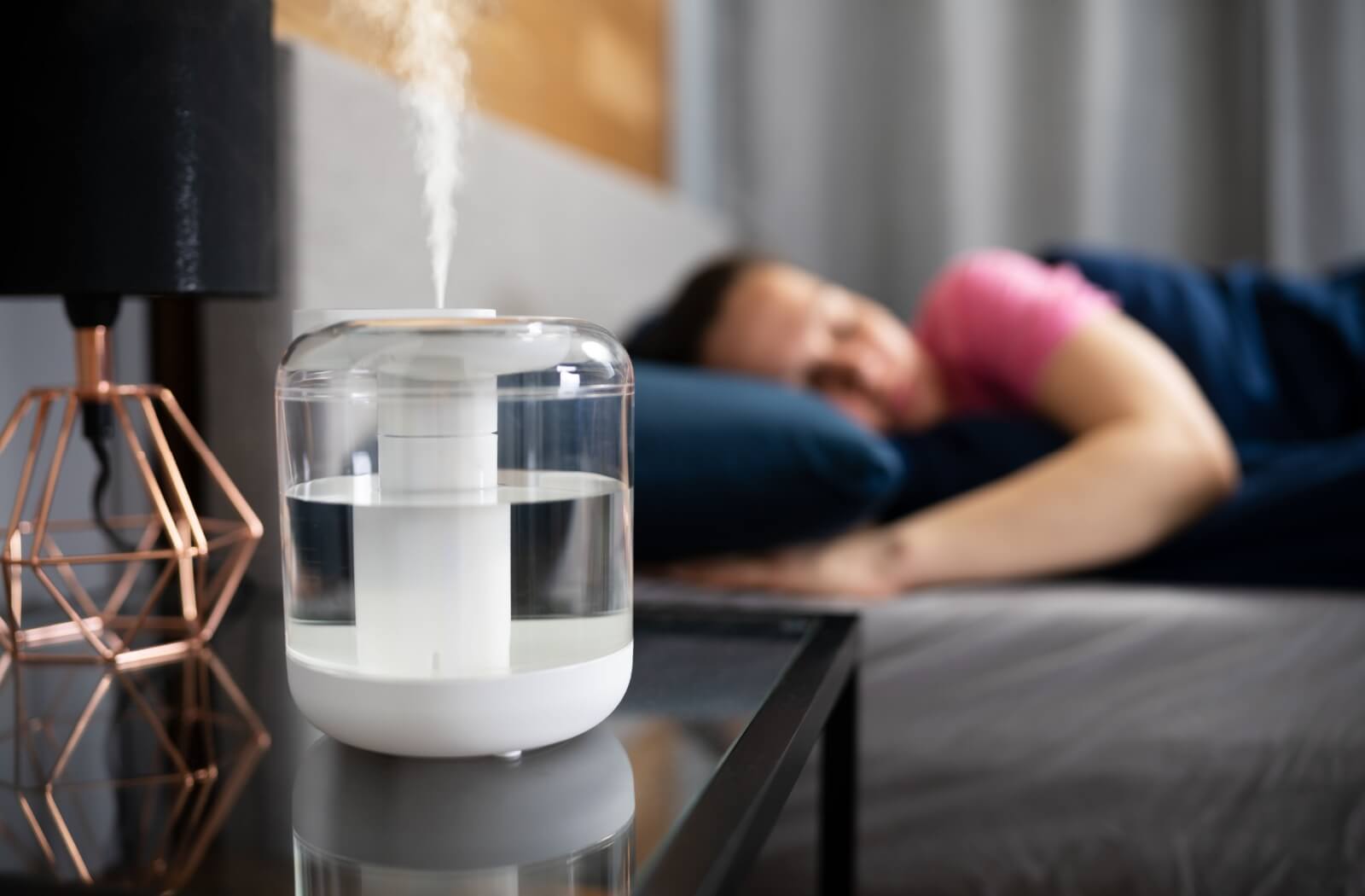 A person in the background sleeping soundly with a humidifier in the foreground to help with moisture in the air to help with dry eyes.